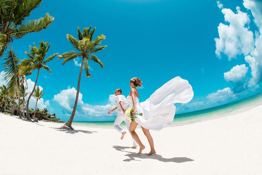 По песчаному побережью бегут счастливые молодожены, держась за руки, жених в белых брюках и рубашке, невеста в белом воздушном - фото 2939781 Свадебное агенство "Travelservice weddings"