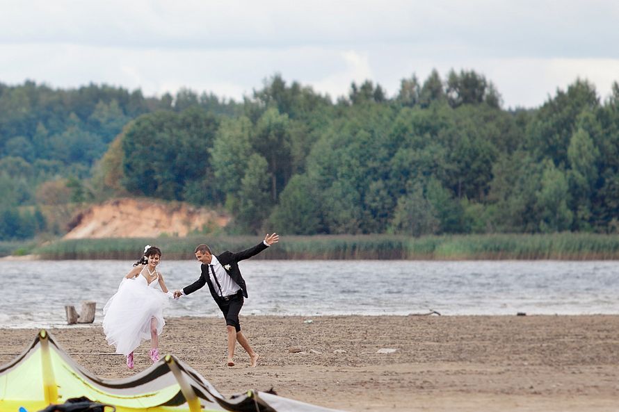По песчаному пляжу бегут счастливые молодожены, жених в черном костюме держит за руку невесту в открытом белом платье, она держит - фото 2318666 Студия АРА - свадебная фотография и видеосъёмка