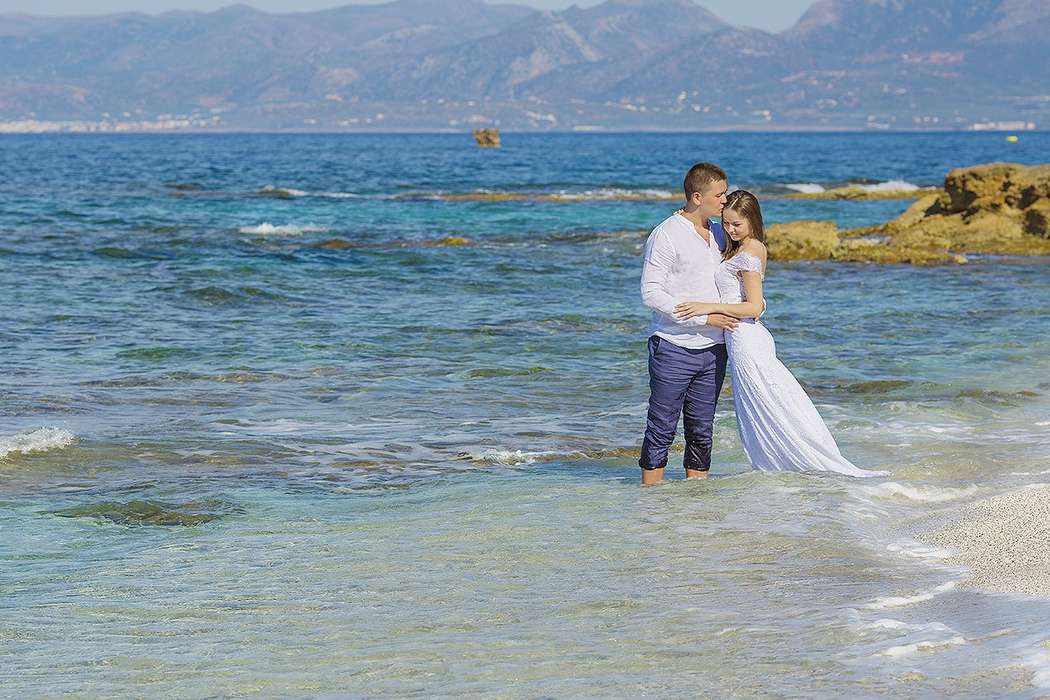 На тропическом пляже, стоя в воде, обнимаются молодожены, жених в синих брюках и белой рубашке шепчет слова любви невесте в белом - фото 2766617 "Tytprod" - фото и видеосъёмка 