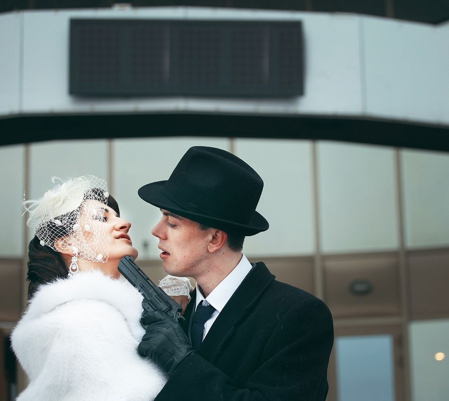 Жених и невеста, прислонившись друг к другу, стоят на фоне здания  с револьвером в руках - фото 3023697 Свадебный фотограф Виктория Романова