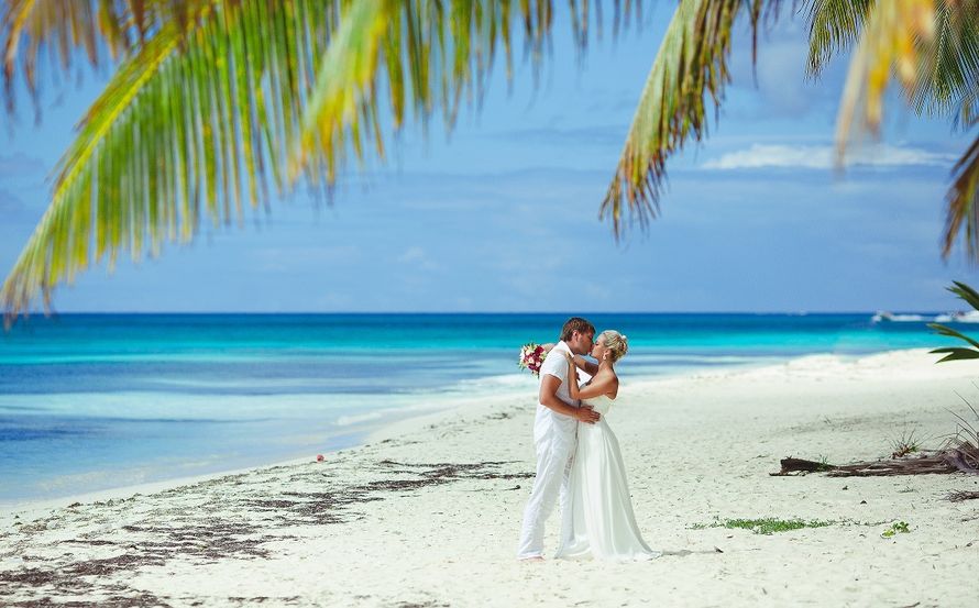 Стоя на пляже, парень в белой шведке и брюках, обняв за талию, целует невесту в белоснежном пышном платье, в руках она держит - фото 3301839 Свадьба в Доминикане от Imagine Wedding