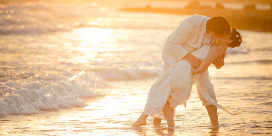Молодожены в белых нарядах целуются стоя босиком в море, жених наклонил свою любимую придерживая ее за талию - фото 614873 webgirl99