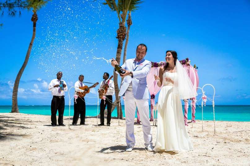 На пляже, стоят жених и невеста, он открывает шампанское, в голубом пиджаке, она держит в руке фужеры, в белом, длинном платье - фото 1252333 Caribbean Wedding - свадьба в Доминикане
