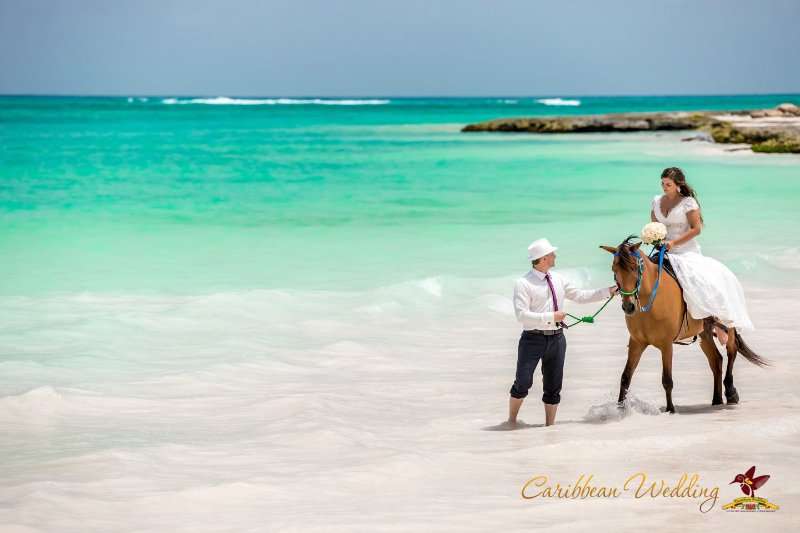 На пляже стоит жених и держит лошадь рукой, невеста сидит на ней в белом, длинном платье с букетом роз - фото 2723967 Caribbean Wedding - свадьба в Доминикане