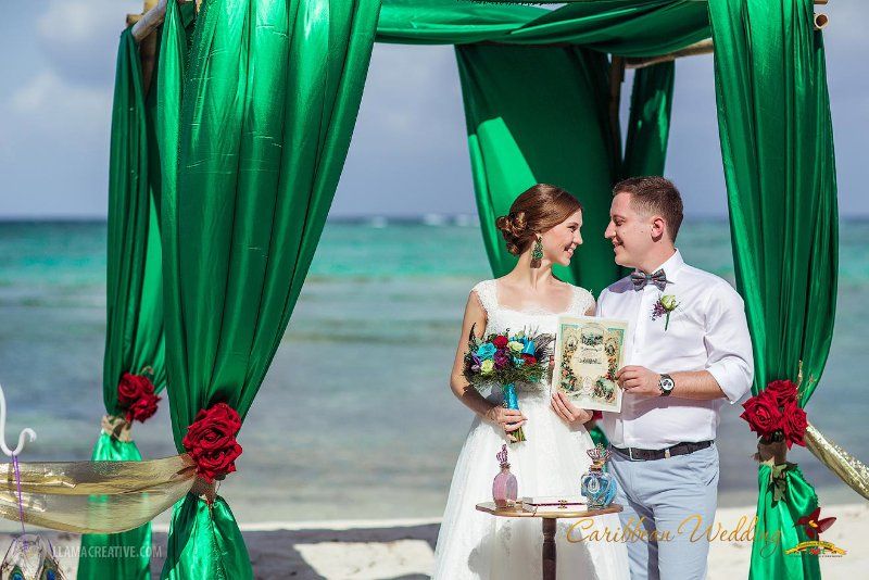 Под сводом изумрудной свадебной арки стоят молодожены, невеста в белом платье держит букет, жених в белой рубашке с бабочкой - фото 3212143 Caribbean Wedding - свадьба в Доминикане